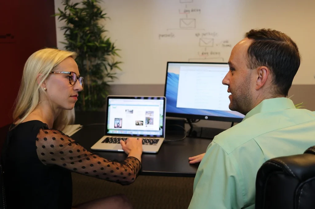 Two colleagues discussing work at a desk with a laptop and monitor, collaborating on a project in a modern office setting.